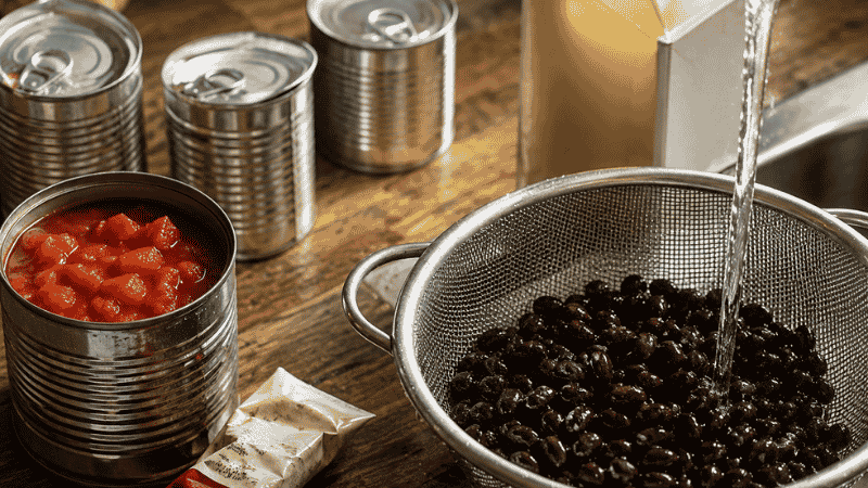 Canned pantry staples on a kitchen counter including black beans being rinsed, diced tomatoes, tomato paste tube, and chicken broth