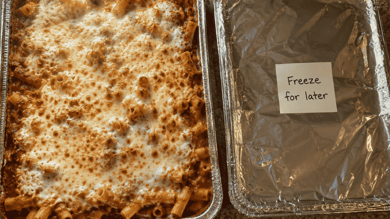 Two pans of baked ziti — one fresh from the oven with bubbling mozzarella, one covered and labeled for freezing — a big family batch cooking strategy for cheap meals