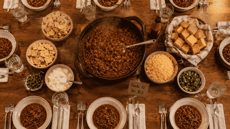 Large pot of beef and bean chili on a family dinner table set for 8 people — a big family meal cheap that feeds 10 for under $12 total