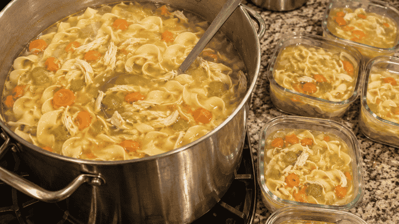 Large stock pot of homemade chicken noodle soup with glass freezer containers being filled beside it — big family batch cooking that feeds 10 to 12 for under $13 total