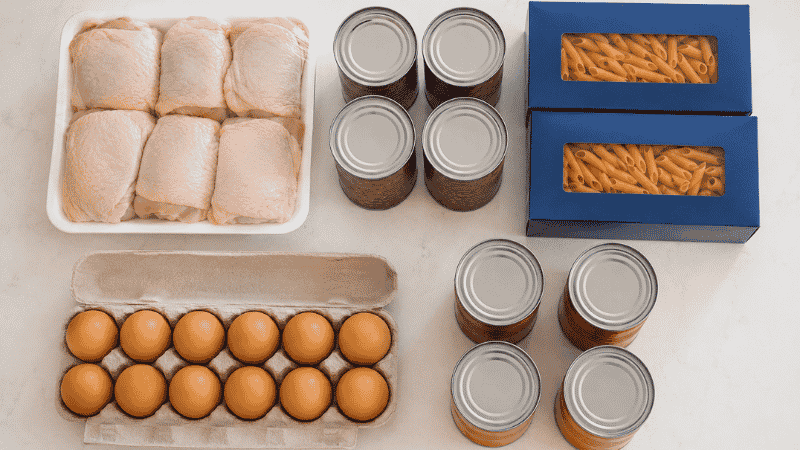 Five budget anchor ingredients for cheap easy family meals: chicken thighs, canned beans, pasta, eggs, and diced tomatoes laid flat on a white kitchen counter