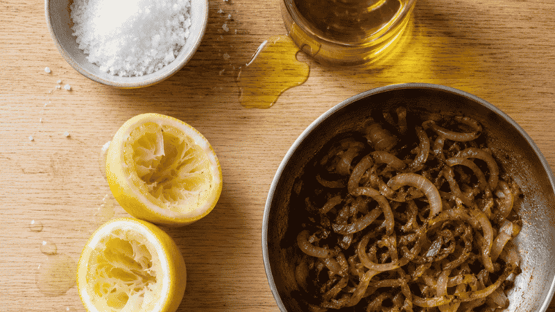 Four flavor-building elements for cheap family meals laid flat: kosher salt, olive oil, lemon, and caramelized onions — the techniques that make budget food taste expensive