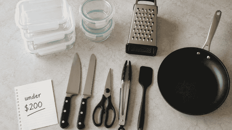 Budget kitchen essentials under $200 laid out flat: non-stick skillet, knives, glass containers, box grater, scissors, tongs, and spatula with a handwritten note