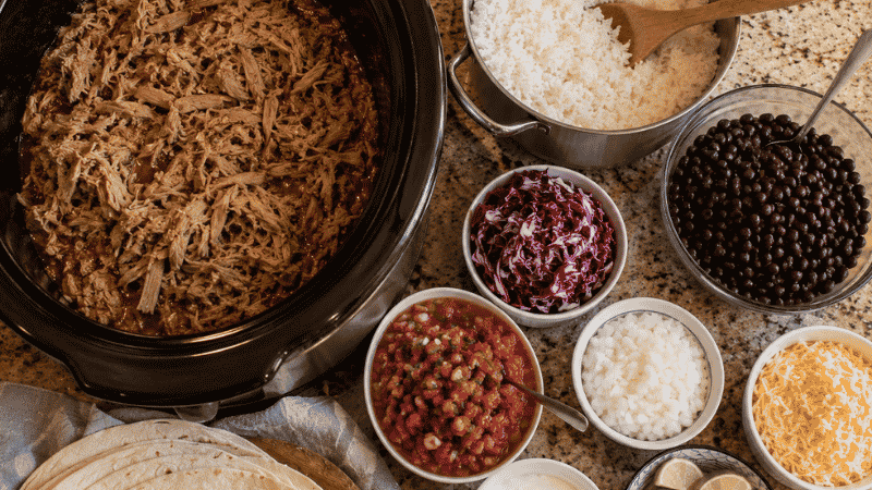 Slow cooker full of shredded pork carnitas beside a build-your-own bowl station with rice, beans, salsa, and toppings — a cheap large family meal that feeds 8 to 10 for under $14