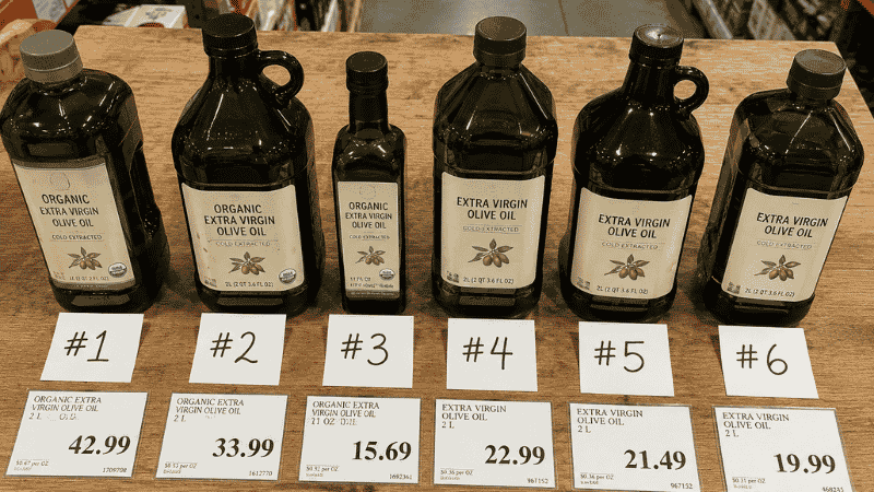 Six Costco olive oil bottles arranged in a row with handwritten ranking cards from number 1 to 6 and price tags — a complete ranked comparison guide for every Costco olive oil option