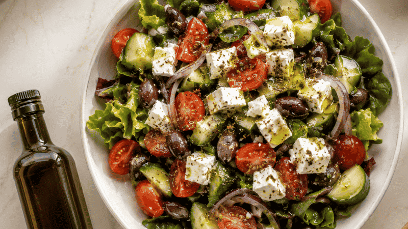 Extra virgin olive oil being drizzled over a colorful Mediterranean salad with olives, tomatoes, and feta — representing the heart-healthy benefits of daily EVOO consumption