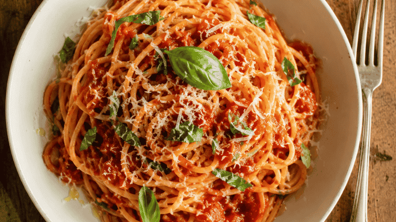 A bowl of perfectly cooked spaghetti in glossy tomato sauce topped with Parmesan and fresh basil — the result of following the correct method for how to boil pasta