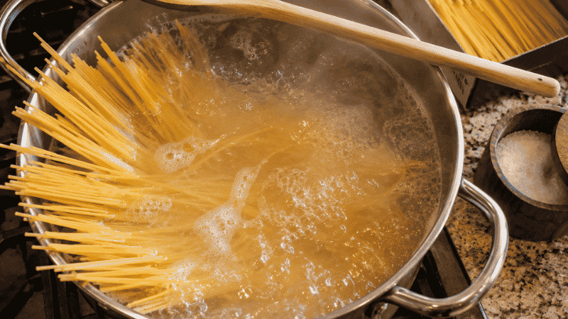 A large pot of vigorously boiling salted water with spaghetti just added — the correct method for how to boil pasta with a wooden spoon resting across the rim