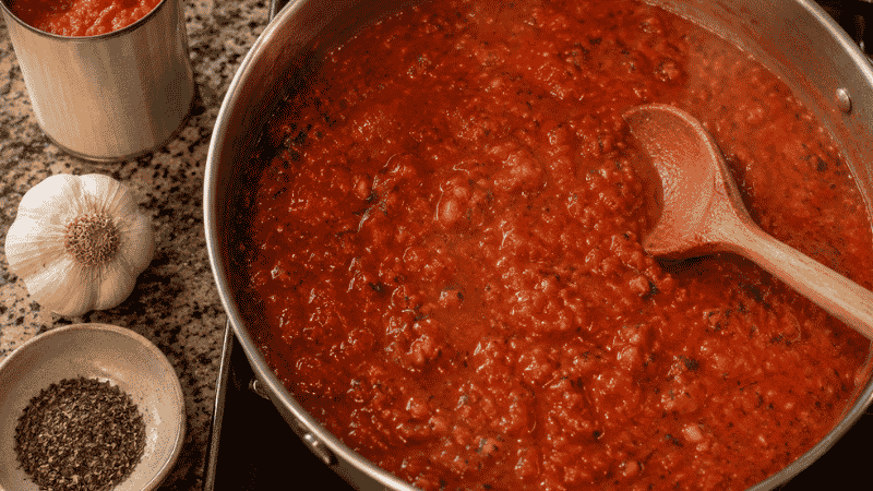 A wide saucepan of rich homemade tomato pasta sauce simmering on the stovetop with a wooden spoon, made from a can of crushed tomatoes, garlic, and dried basil