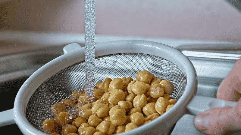 A close-up of white chickpeas being rinsed under a kitchen faucet in a stainless steel colander to remove sodium and preservatives.
