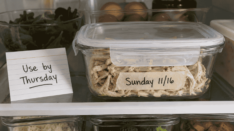 Glass airtight container of cooked shredded chicken labeled with Sunday date and use by Thursday notecard in a refrigerator shelf — how long does cooked chicken last in the fridge guide