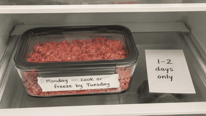 Raw ground beef in a sealed airtight container labeled Monday with a cook or freeze by Tuesday note on a refrigerator bottom shelf — how long does ground beef last in the fridge guide showing the 1 to 2 day storage rule