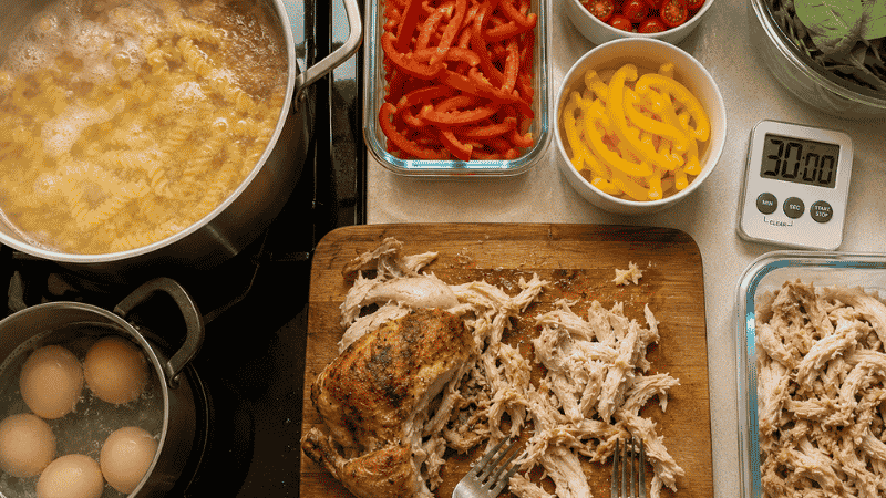 Sunday meal prep session in progress showing pasta boiling, eggs cooking, rotisserie chicken being shredded, and vegetables already prepped with a 30-minute kitchen timer — the complete Sunday prep schedule for a week of lunches