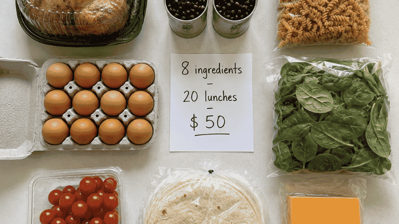 Eight budget anchor grocery ingredients laid flat including rotisserie chicken, canned beans, pasta, eggs, spinach, cherry tomatoes, tortillas, and cheese with a $50 for 20 lunches notecard
