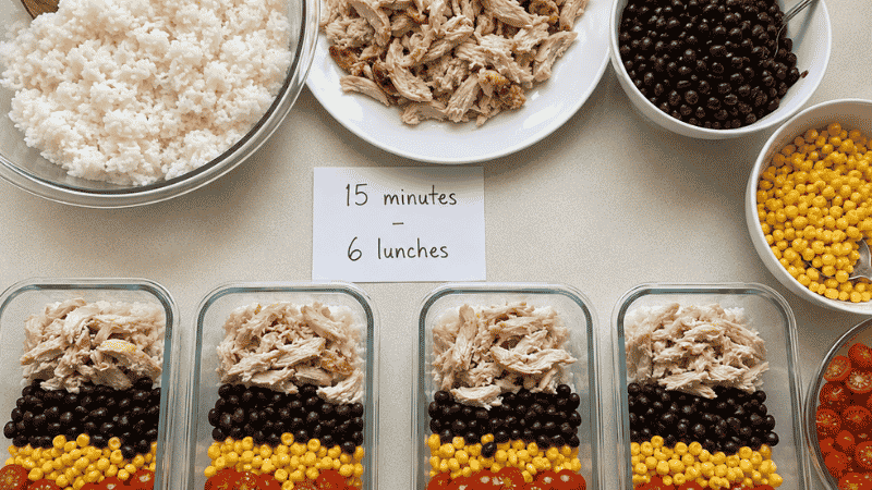 Meal prep assembly line with shredded rotisserie chicken, cooked rice, beans, corn, and cherry tomatoes being divided into four glass containers — 15 minutes of prep unlocking 6 different lunch ideas
